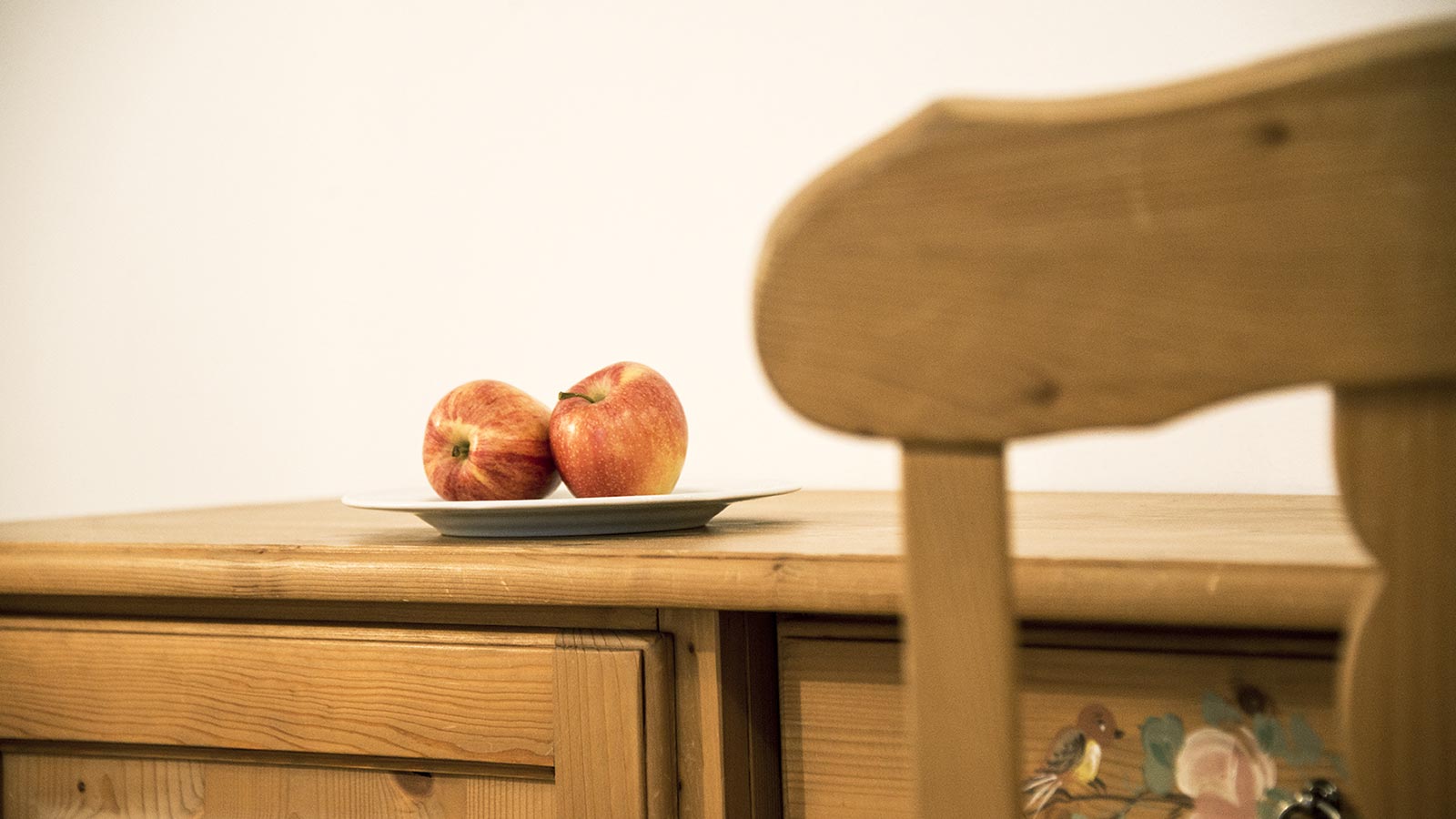 Cassapanca in legno, sulla quale è appoggiato un piatto con due mele rosse, e schienale sfocato di una sedia in legno